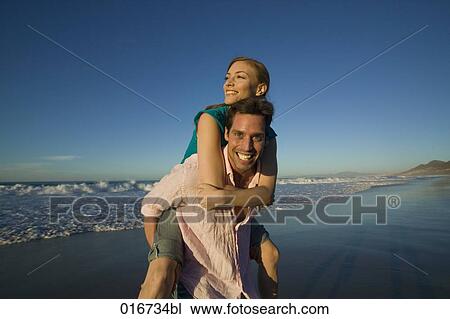 Stock Image - young man giving woman piggyback ride on beach. Fotosearch