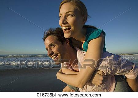 Stock Image - young man giving woman piggyback ride on beach. Fotosearch