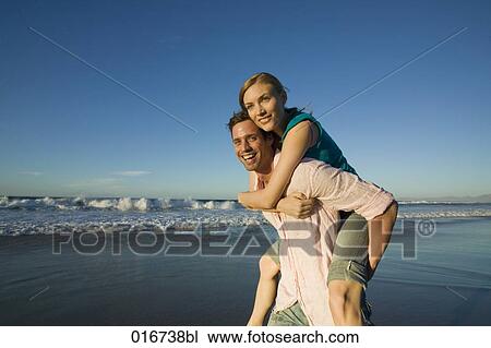 Stock Image - young man giving woman piggyback ride on beach. Fotosearch