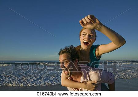 Stock Image - young man giving woman piggyback ride on beach. Fotosearch