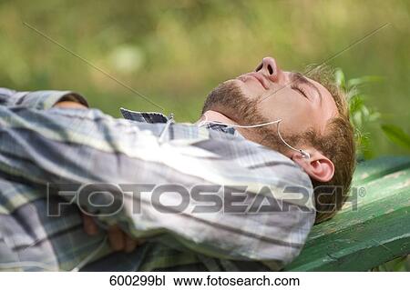Stock Image - young man lying on park bench wearing earphones listening to music. Fotosearch