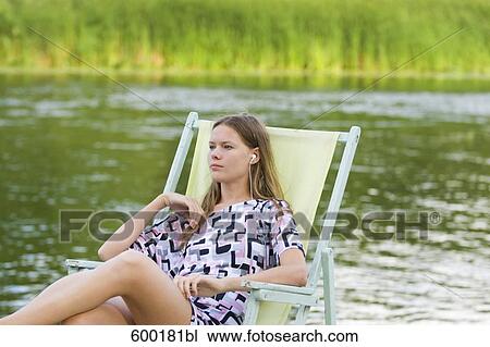 Stock Image - young woman sitting in sunlounger listening to music on mp3 player. Fotosearch