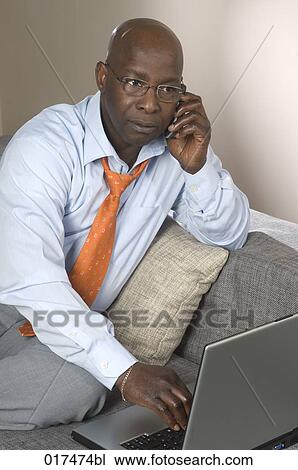 black businessman sitting on sofa with laptop listening to mobile phone View Large Photo Image Stock Image - black businessman sitting on sofa with laptop listening to mobile phone. Fotosearch