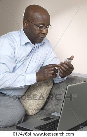 Stock Image - black businessman sitting on sofa with laptop checking his mobile phone for text messages. Fotosearch