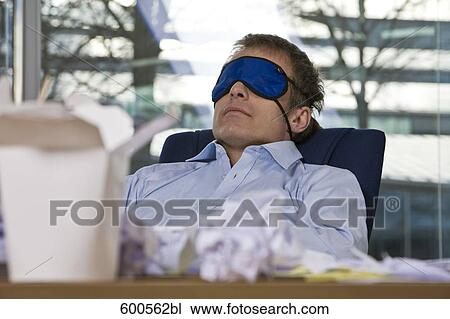 Stock Image - businessman taking a nap at his desk wearing a sleep mask. Fotosearch