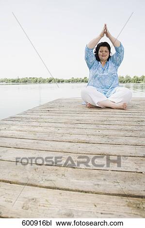 Stock Image - overweight woman meditating on pier by lake. Fotosearch