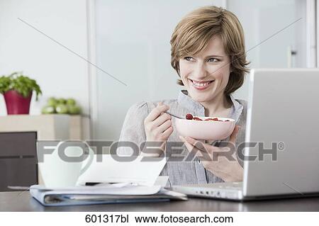 Stock Image - portrait of woman at home in front of computer eating snack. Fotosearch