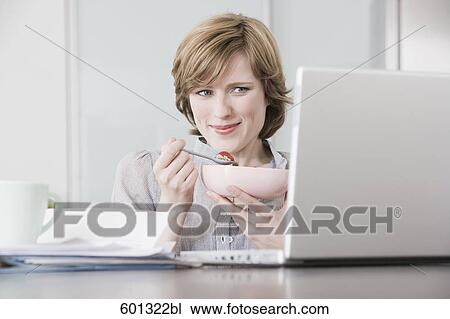 Stock Image - portrait of woman at home in front of computer eating snack. Fotosearch