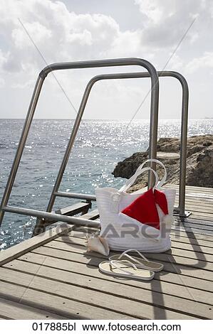 Stock Image - still life of bag flip flops and santa claus hat on pier. Fotosearch