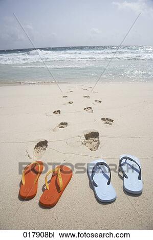 Stock Image - two pairs of flip flops and footsteps on sandy beach. Fotosearch
