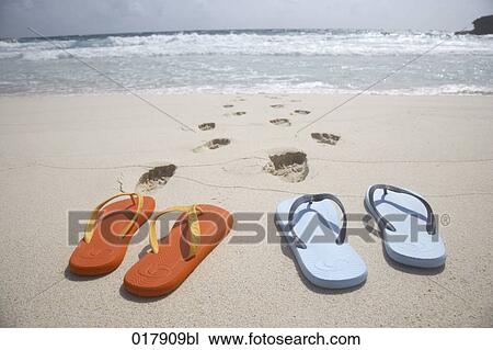 Stock Image - two pairs of flip flops and footsteps on sandy beach. Fotosearch