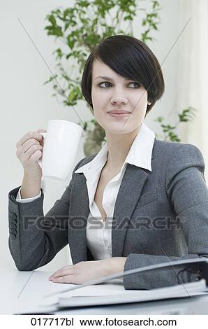 Stock Image - young businesswoman sitting at desk holding cup. Fotosearch