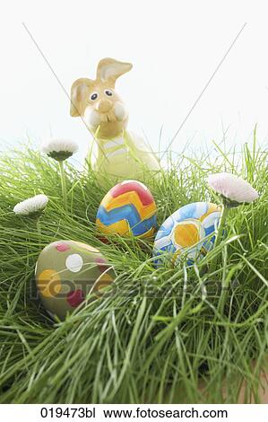 Stock Image - figure of bunny and three painted easter eggs lying in grass. Fotosearch