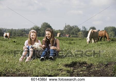 Stock Image - full body portrait of two girls sitting on meadow with dog at horse ranch. Fotosearch