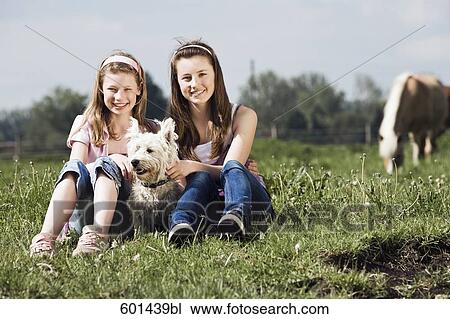 Stock Image - full body portrait of two girls sitting on meadow with dog at horse ranch. Fotosearch
