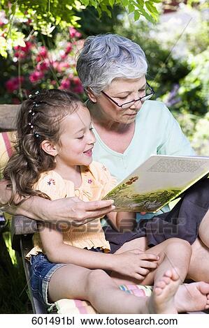 Stock Image - grandmother sitting in garden reading from book to little girl. Fotosearch