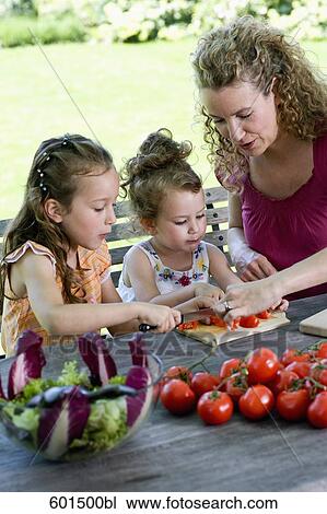 Stock Image - mother showing her two daughters how to make salad of tomatoes. Fotosearch