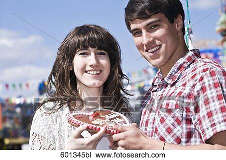 Stock Image - portrait of teenage couple with gingerbread heart at fun fair. Fotosearch