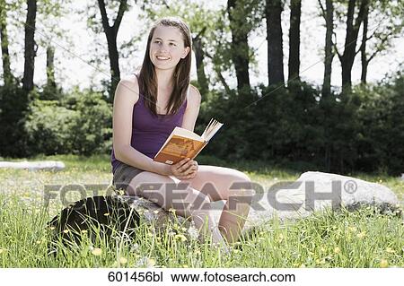 Stock Image - portrait of young girl sitting on tree trunk reading book. Fotosearch