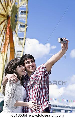 Stock Image - portrait teenage couple taking photo of themselves at fun fair. Fotosearch