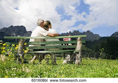 Stock Image - rear view of mature couple sitting on bench in front of mountains. Fotosearch