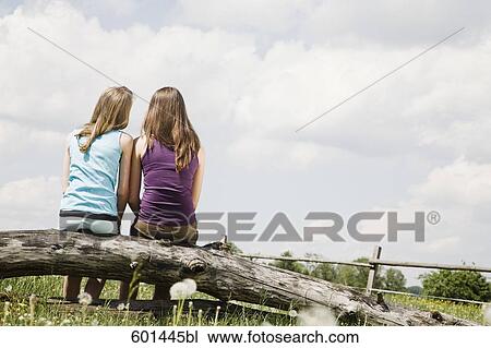 Stock Image - rear view of two girls sitting on tree trunk looking at sky. Fotosearch
