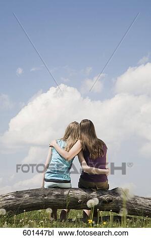 Stock Image - rear view of two girls sitting on tree trunk embracing each other. Fotosearch