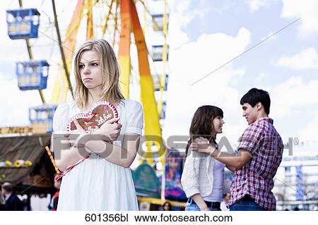 Stock Image - sulking teenage girl at fun fair with couple in background. Fotosearch