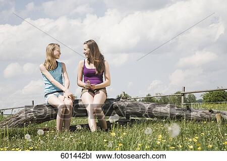 Stock Image - two girls sitting together on tree trunk at horse ranch. Fotosearch