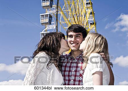 Stock Image - two teenage girls kissing boy on cheek at fun fair. Fotosearch