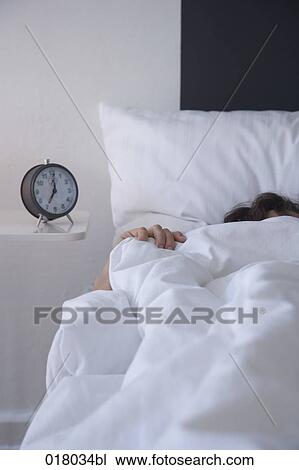 Stock Image - woman in bed with alarm clock on sideboard. Fotosearch