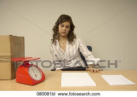 Stock Image - bored female office worker sitting at desk looking at scales and package on desk. Fotosearch