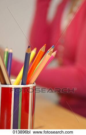 Stock Image - cup with colourful pencils on table, woman in background. Fotosearch
