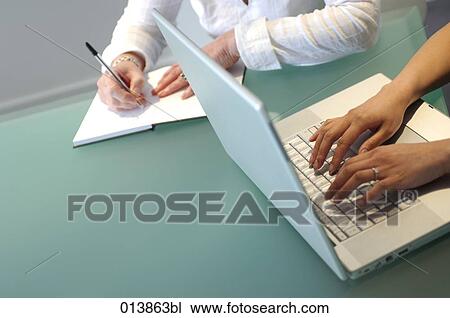 Stock Image - detail of one woman typing on laptop and the other writing in diary. Fotosearch