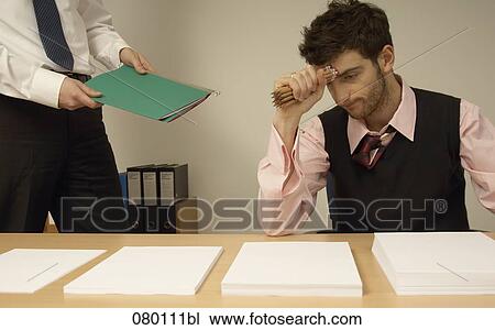 Stock Image - frustrated businessman sitting at desk full of documents colleague giving him file. Fotosearch