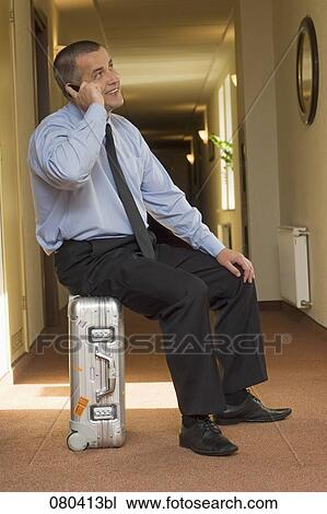 Stock Image - full body portrait of mature businessman sitting on suitcase in hotel corridor and talking on mobile phone. Fotosearch