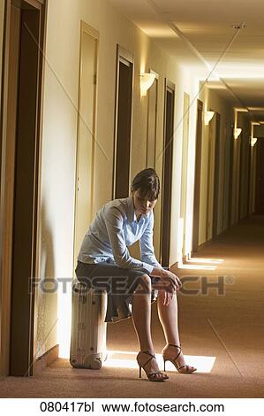 Stock Image - full body portrait of young businesswoman sitting on suitcase in hotel corridor. Fotosearch