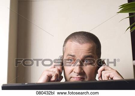 Stock Image - headshot of mature businessman listening to two mobile phones at once and looking at screen of laptop computer. Fotosearch