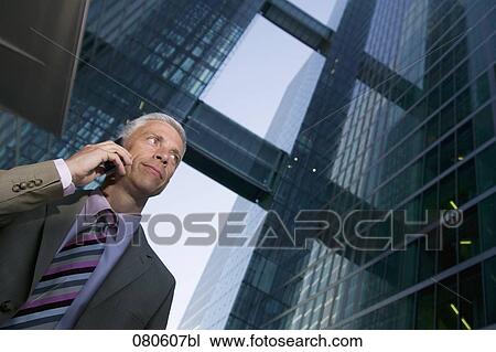 Stock Image - low angle portrait of mature businessman talking on mobile phone in front of office building. Fotosearch