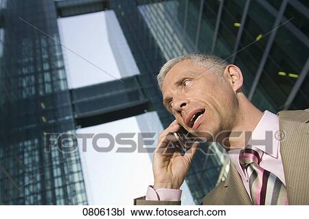 Stock Image - low angle portrait of mature businessman talking on mobile phone in front of office building. Fotosearch