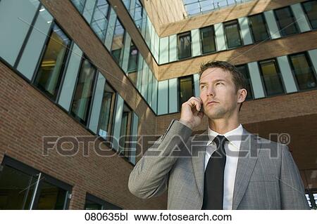Stock Image - low angle view of businessman listening to mobile phone in front of office building. Fotosearch