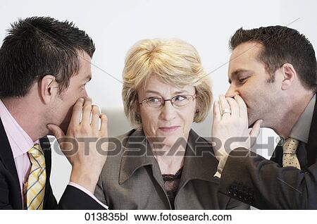 Stock Image - mature businesswomen listening to two male colleagues whispering. Fotosearch