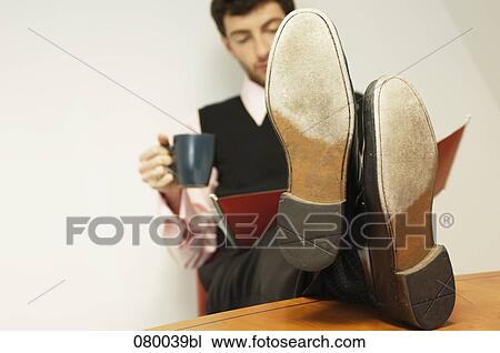 Stock Image - portrait of businessman sitting at table with his feet up holding mug and reading file. Fotosearch