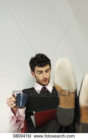 Stock Image - portrait of businessman sitting at table with his feet up holding mug and reading file. Fotosearch