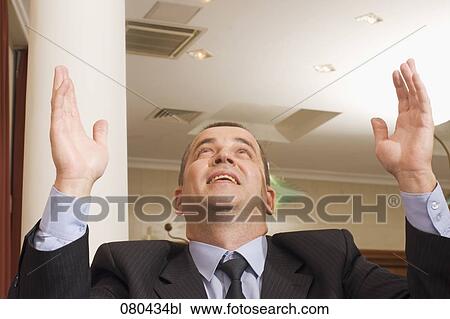 Stock Image - portrait of businessman sitting in hotel lobby raising his hands and looking up. Fotosearch