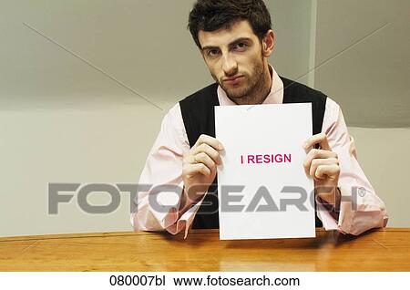 Stock Image - portrait of frustrated businessman sitting at desk holding resign document. Fotosearch