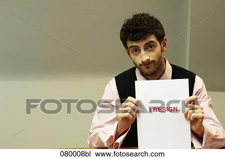 Stock Image - portrait of frustrated businessman sitting at desk holding resign document. Fotosearch