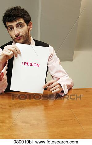 Stock Image - portrait of frustrated businessman sitting at desk holding resign document. Fotosearch