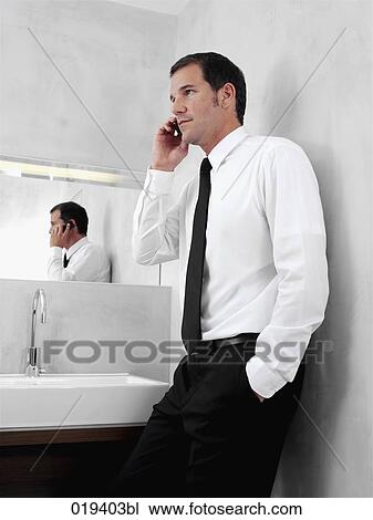 Stock Image - portrait of young businessman in front of mirror in bathroom talking on mobile phone. Fotosearch