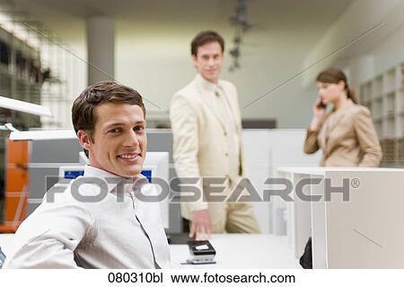 portrait of young office worker sitting at desk with colleagues in the background View Large Photo Image Stock Image - portrait of young office worker sitting at desk with colleagues in the background. Fotosearch
